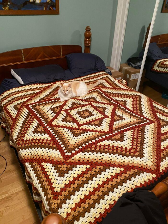 A fluffy white and orange cat rests on a large, intricately patterned DIY crochet blanket in shades of brown, red, and cream.