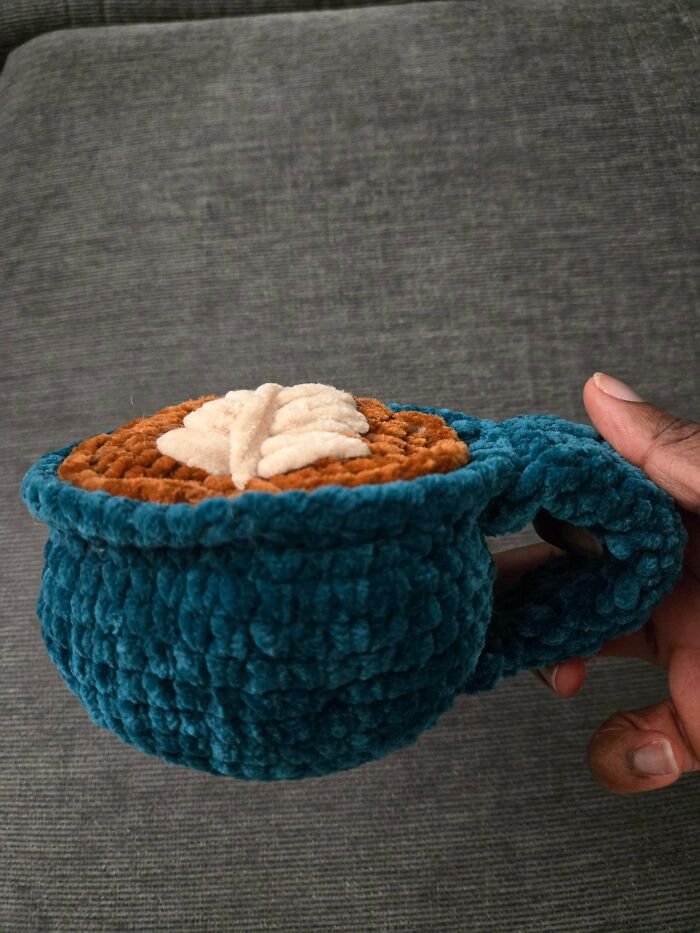 A hand holds a charming DIY crochet mug, teal in color, with a brown top and a white leaf design.
