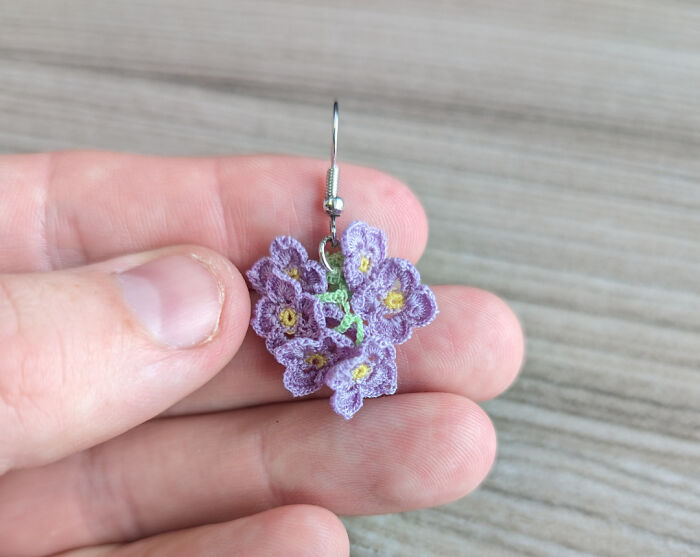 A hand holds a delicate DIY crochet earring shaped like a cluster of tiny purple flowers with yellow centers.