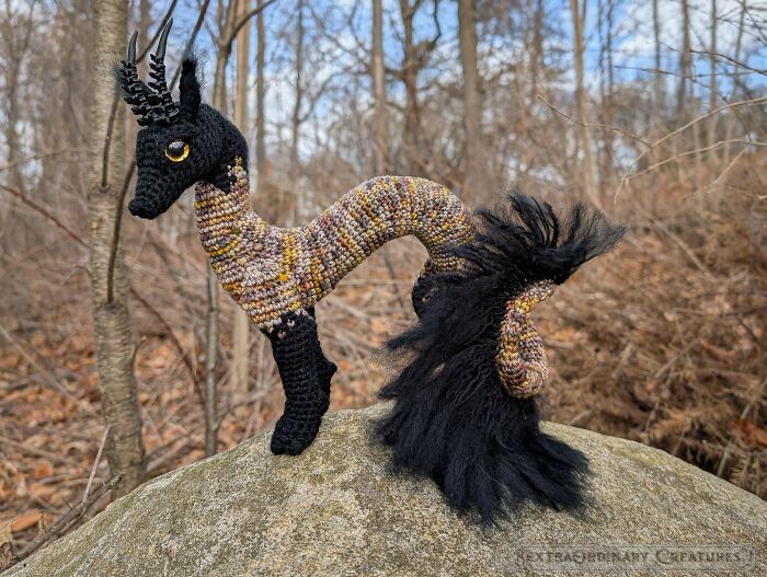 A DIY crochet creature with black legs and head, a yellow eye, and a long, patterned body with a fluffy black tail.