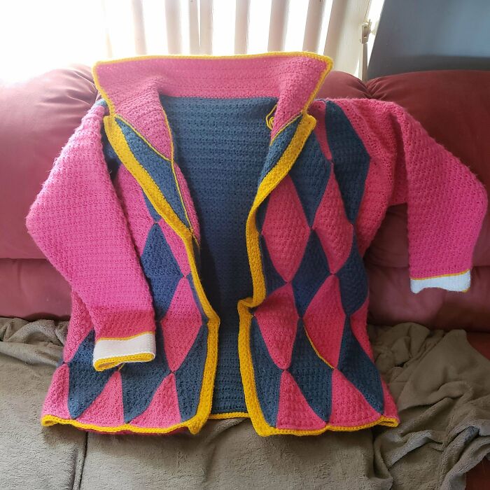 A pink and blue diamond-patterned DIY crochet jacket with yellow trim, laid out on a couch.