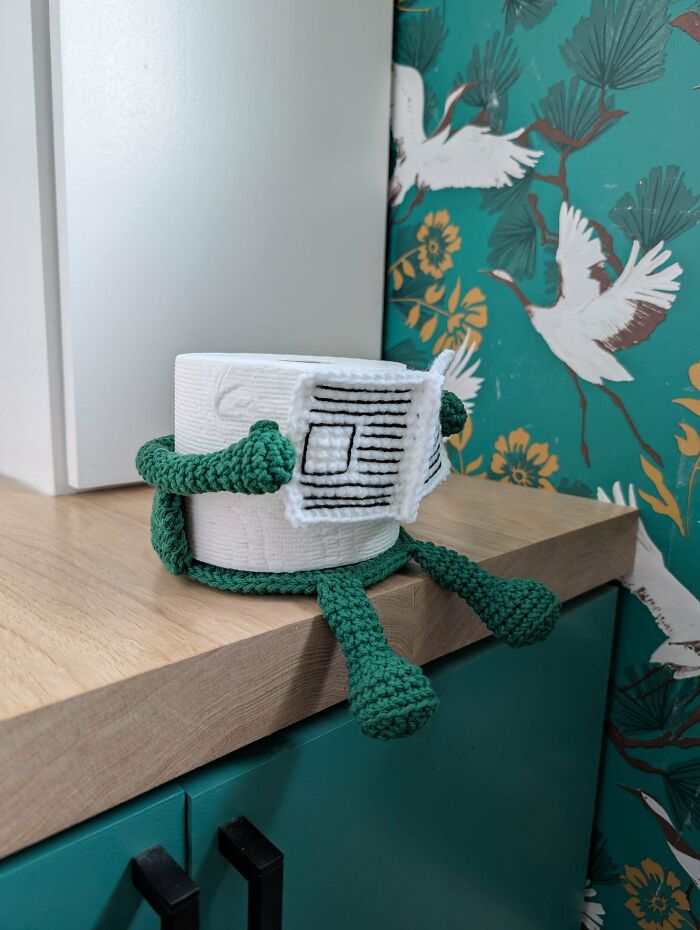 A toilet paper roll with a green DIY crochet cover is reading a tiny newspaper, a useful DIY creation.
