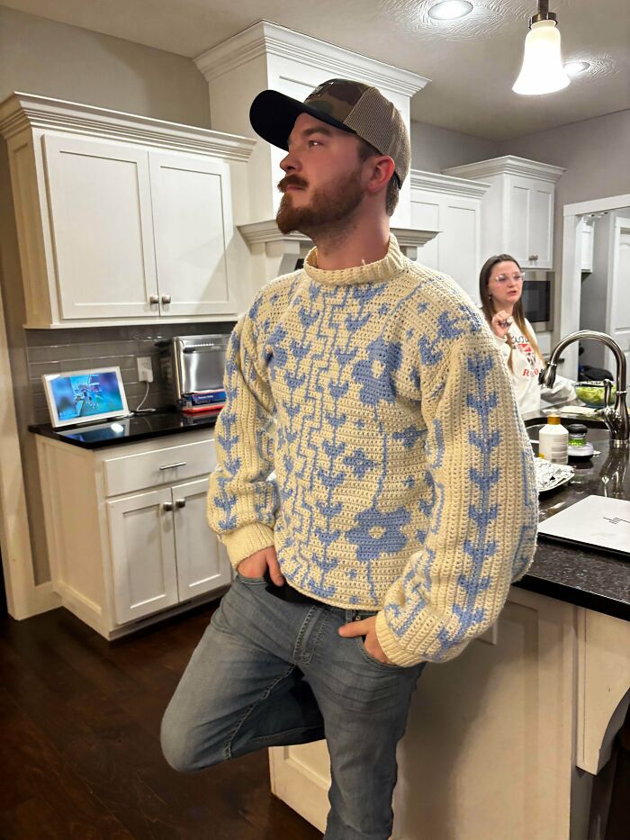 A man wearing a blue and white patterned DIY crochet sweater stands in a kitchen, showing off a useful crochet creation.
