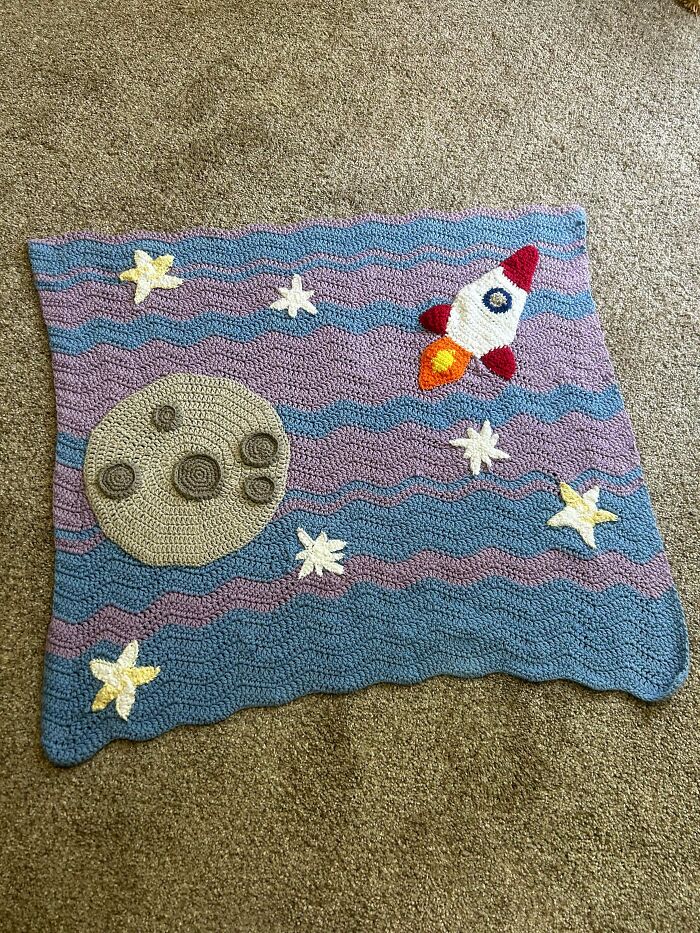 A crochet blanket featuring a rocket, moon, and stars against a wavy blue and purple background, a useful DIY creation.