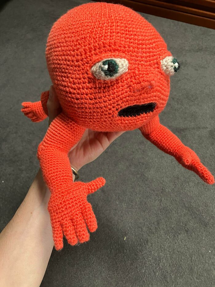 A hand holds a peculiar, red, useful DIY crochet creature with wide eyes.
