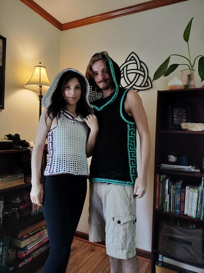 A couple wearing their most useful DIY crochet tops, a white hooded top for her and a black hooded vest for him, in a living room.