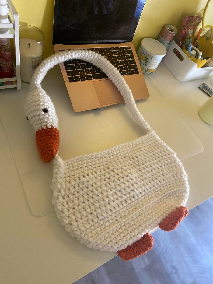 A white crochet goose-shaped bag with orange accents, a unique and useful DIY creation, on a desk next to a laptop.
