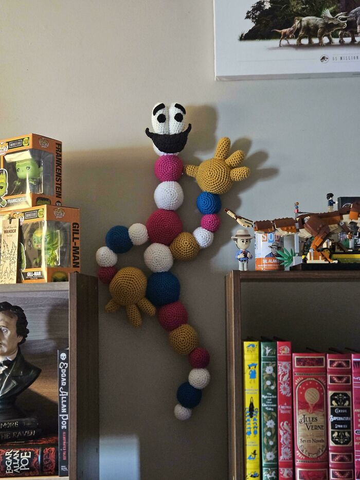 A colorful, segmented crochet figure, a creative DIY creation, mounted on a wall above a bookshelf with collectibles.