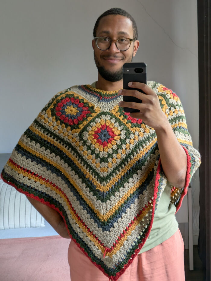A man in a mirror selfie, wearing a stylish, multi-colored crochet poncho, a useful DIY creation.