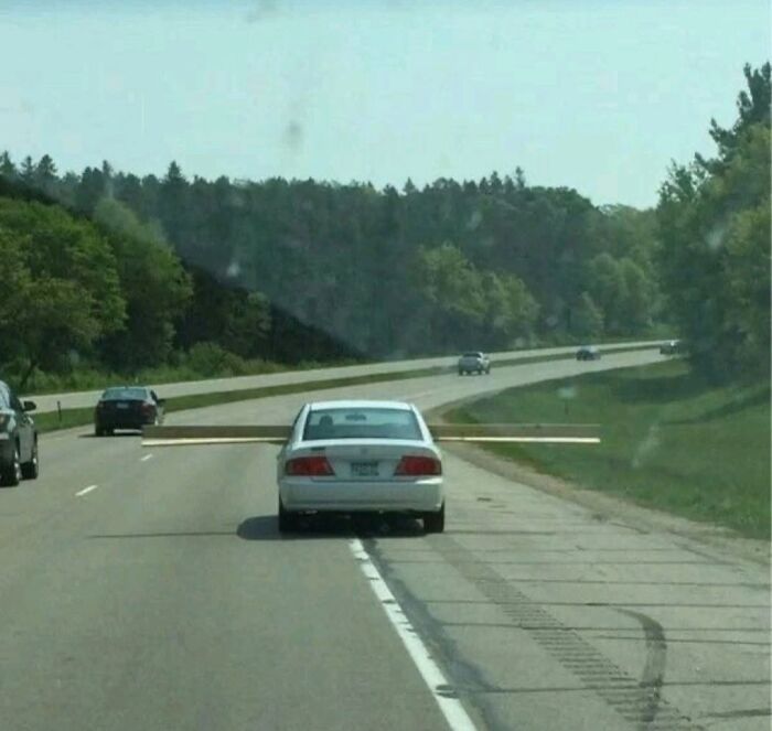 A white car driving down a highway with a very long piece of wood sticking out of its trunk, a ridiculous post.