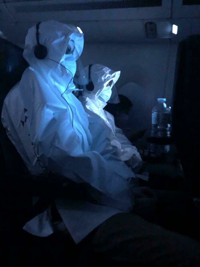 Two figures in white full-body protective suits and headphones sit in a dark airplane cabin, illuminated by blue light, a creepy image for nightmare fuel.