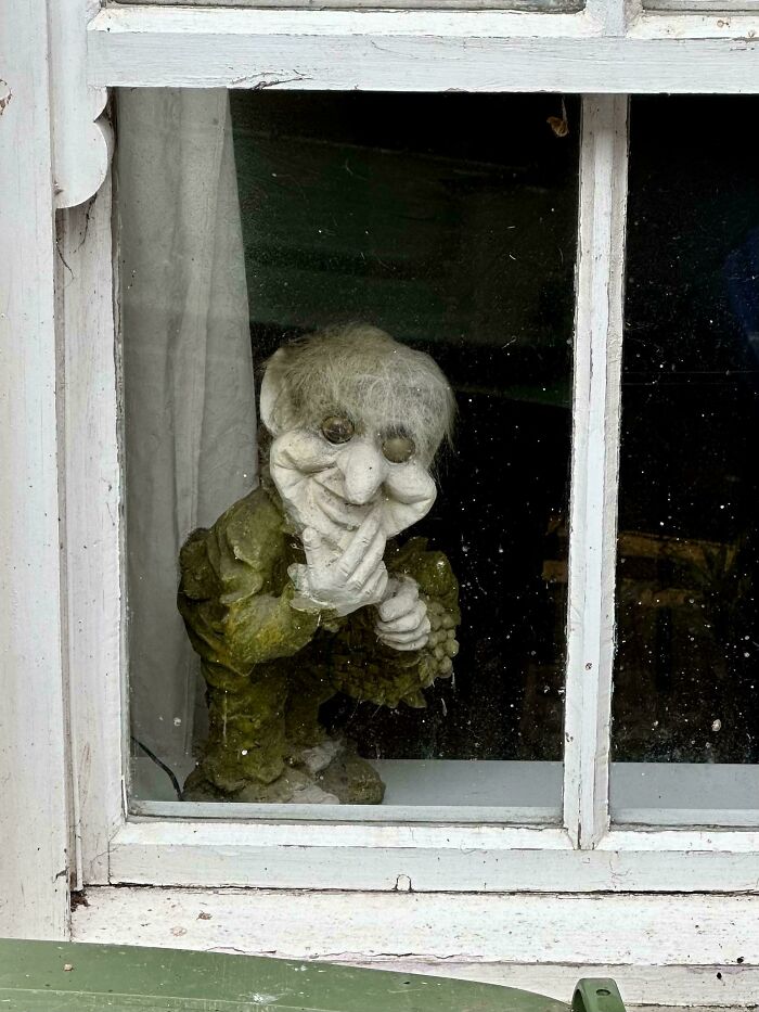 A creepy, gnarled goblin-like figure with white hair peeking through a dirty window, truly nightmare fuel.
