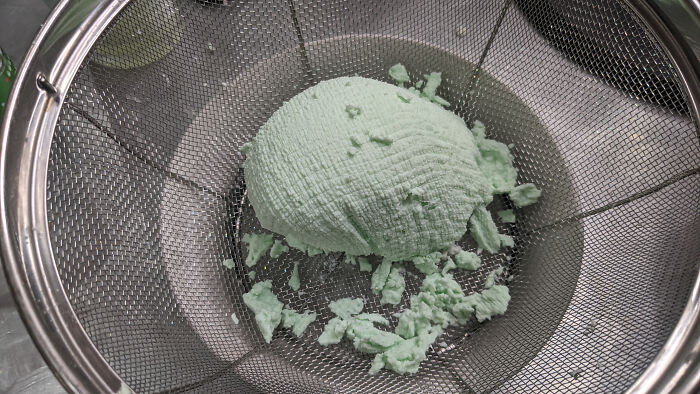 A pale green, crumbly mass in a metal colander, resembling cottage cheese or dough, depicting a piece of disgusting food.