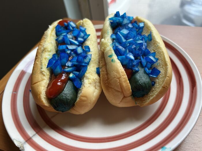 Two hot dogs with blue buns and blue relish, ketchup, and what appears to be a greenish sausage, presenting as truly disgusting food.