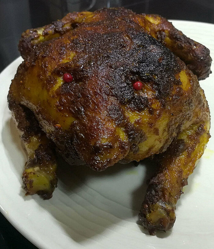 A whole roasted chicken, possibly overcooked, with a dark, almost burnt crust and red peppercorns for eyes, appearing as disgusting food.