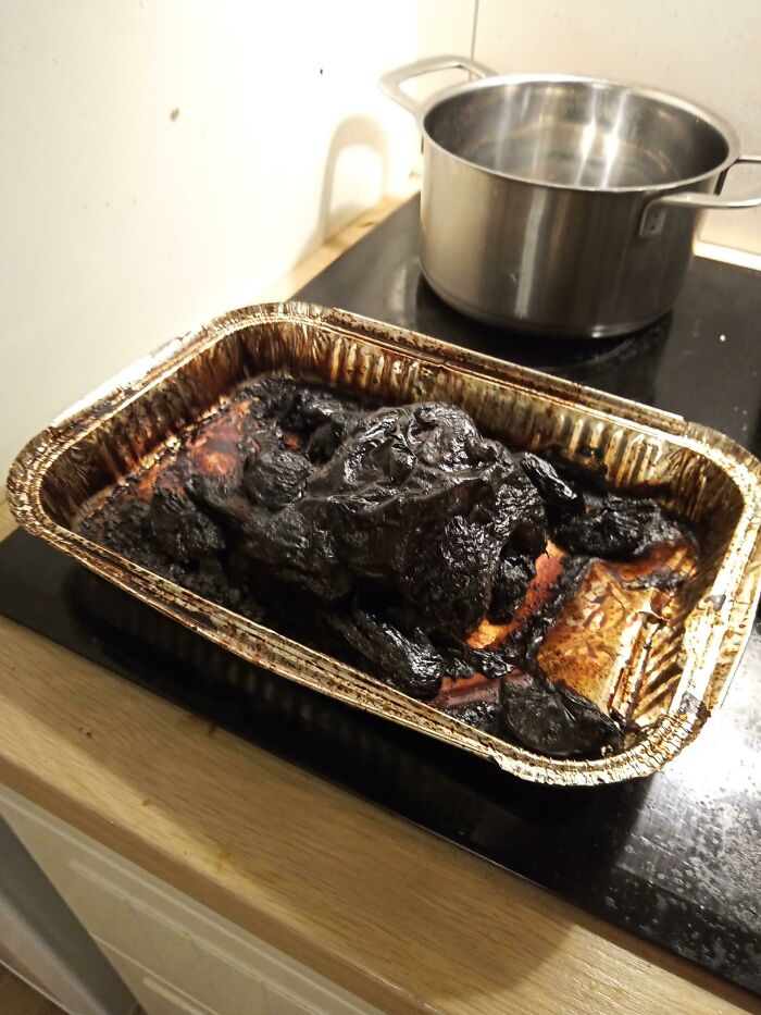 A severely burnt and blackened piece of disgusting food in a foil baking pan on a stovetop next to a metal pot.