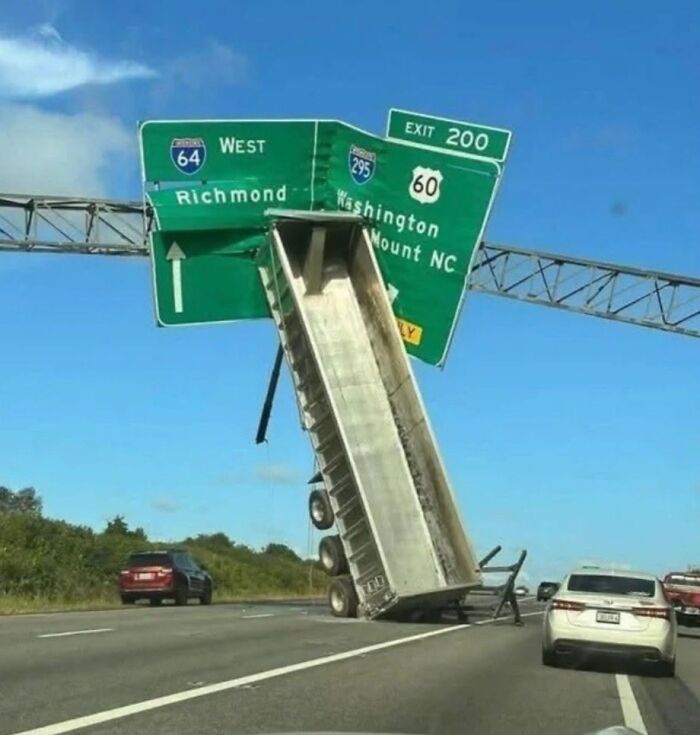 A dump truck with its bed raised, caught under an interstate sign, a ridiculous post showing cultural decline.