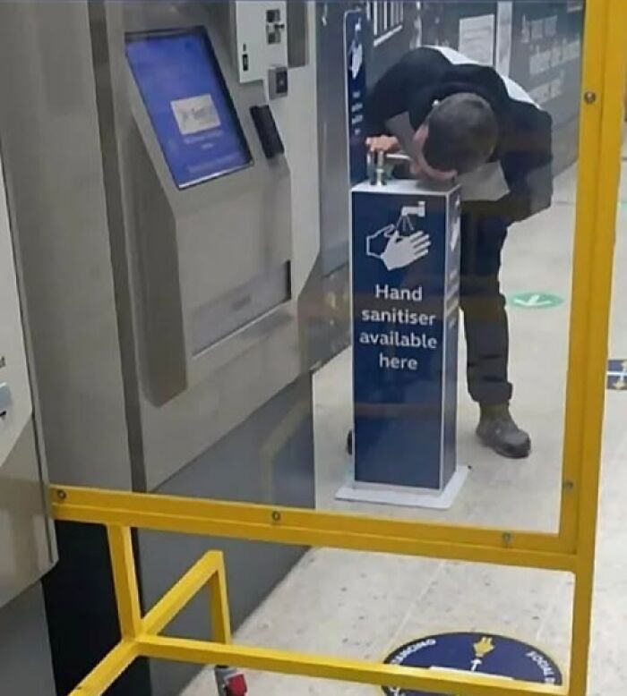 A person drinking from a hand sanitizer dispenser, highlighting a cultural decline into idiocy.