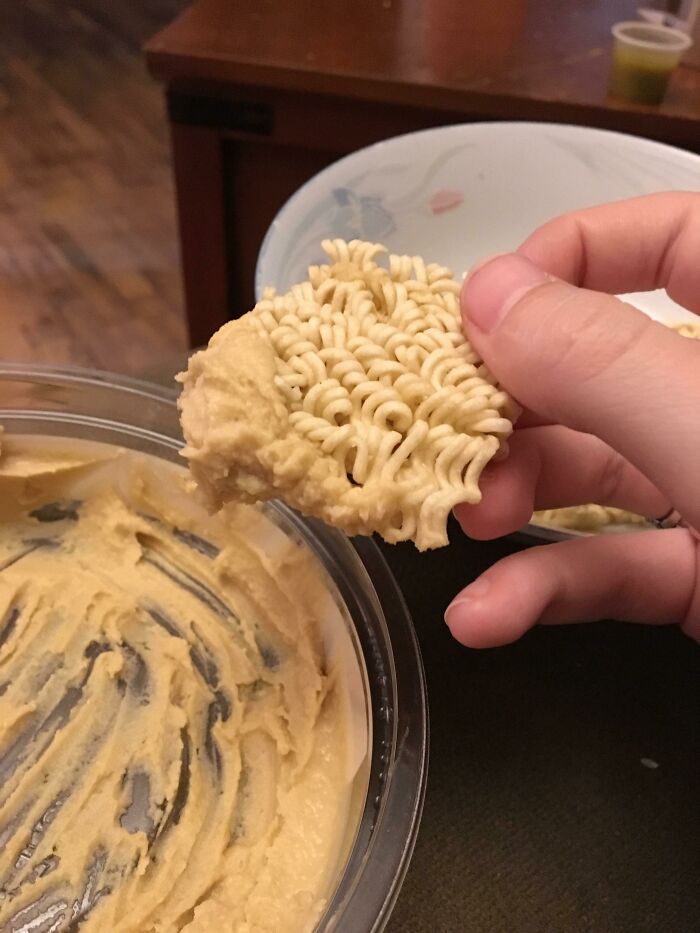 A hand holding a piece of dry ramen noodles dipped in hummus, an example of stupid vegan food.