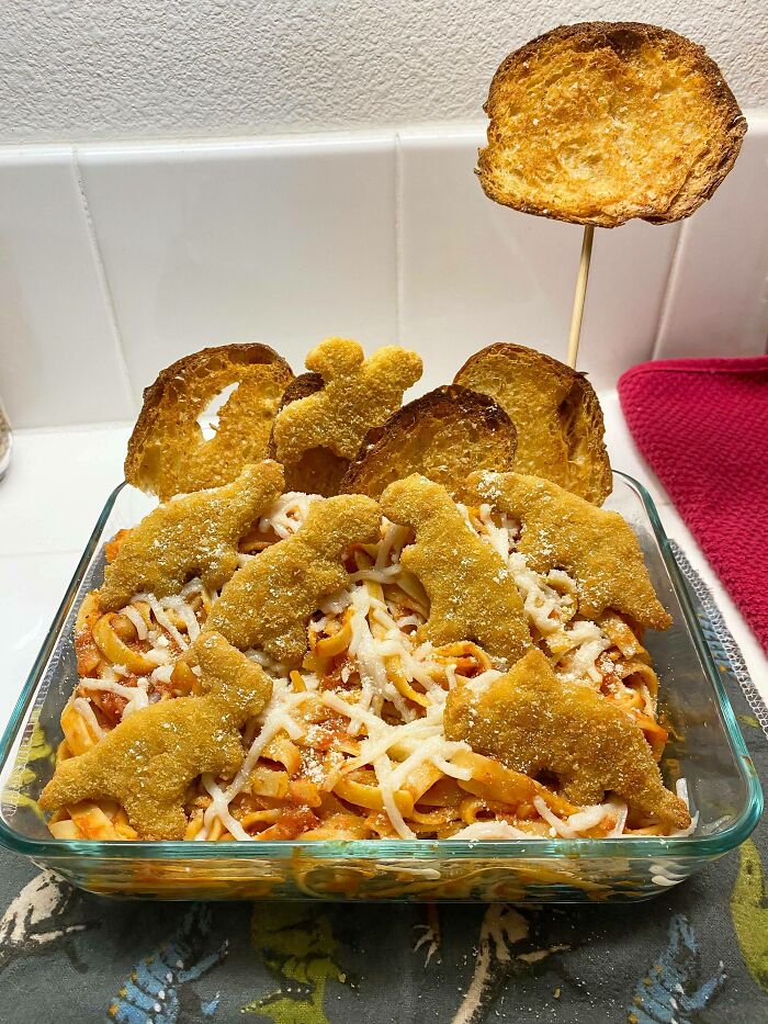 A glass baking dish of pasta topped with dinosaur-shaped nuggets and cheese, a playful take on vegan food.