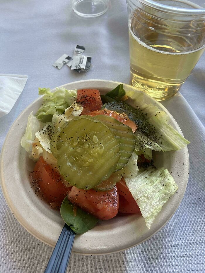 A small bowl of vegan food salad with lettuce, tomatoes, and large pickle slices, next to a drink.