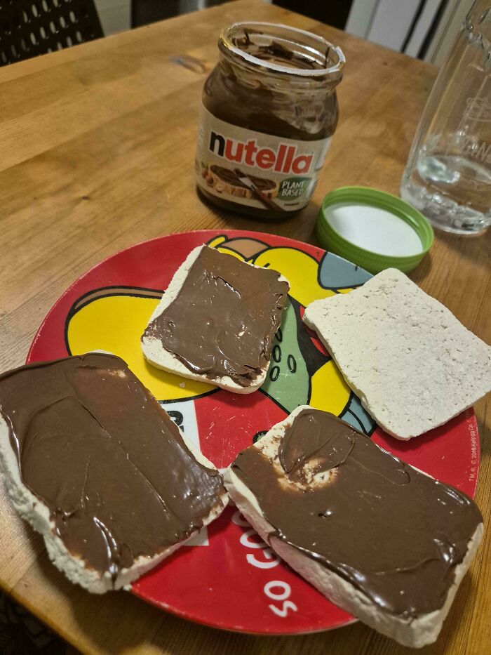 An open jar of plant-based Nutella sits behind four pieces of bread spread with chocolate, representing vegan food.