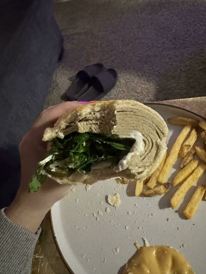 A hand holds a half-eaten vegan food sandwich with layers of light-colored filling and green lettuce, next to fries.