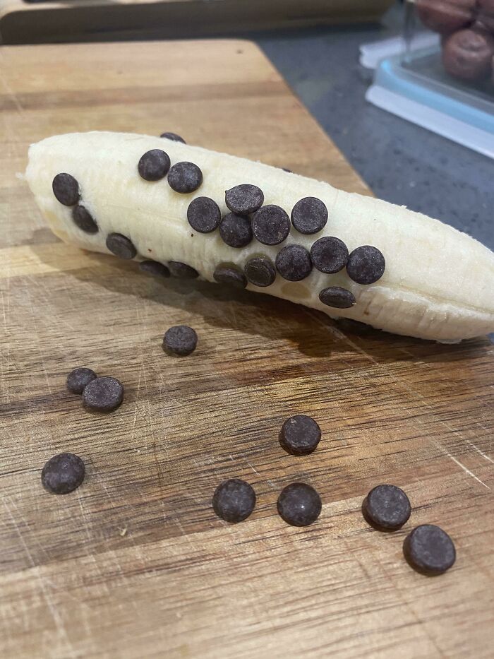 A peeled banana with scattered chocolate chips on a wooden cutting board, an example of stupid vegan food.