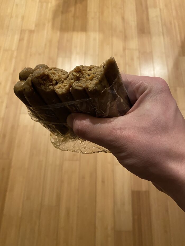 A hand holding a package of brown, sausage-like vegan food, appearing somewhat unappetizing.