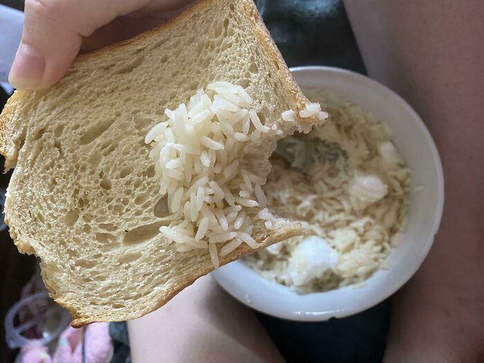 A hand holding a slice of bread with plain white rice piled on top, beside a bowl of soupy rice, a piece of stupid vegan food.