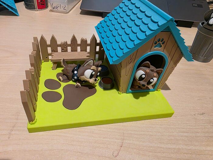 A small doghouse diorama with two cartoonish puppies, a fence, and a bench, demonstrating useful things people printed with their 3D printers.