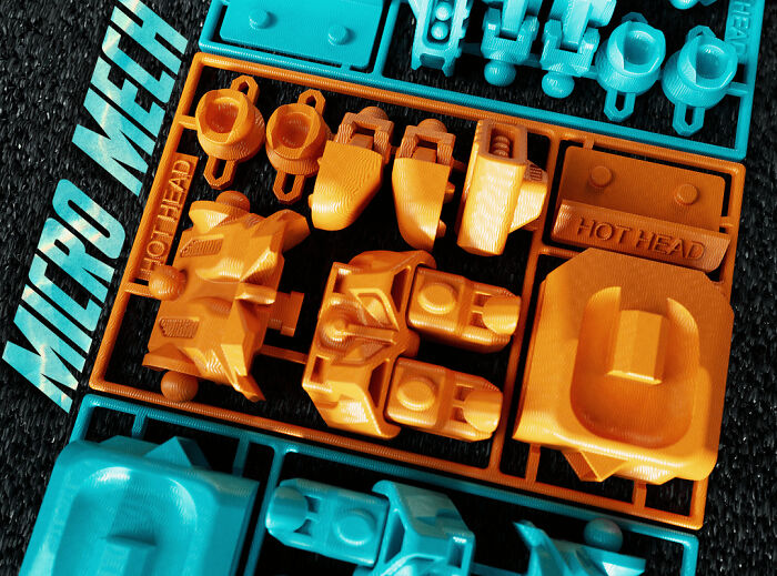 A close-up of orange and blue plastic parts, still attached to their frames, printed with a 3D printer.