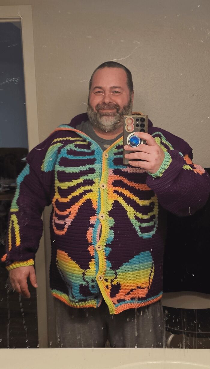 A man smiles in a mirror, showcasing his colorful skeleton crochet cardigan, a useful DIY creation.
