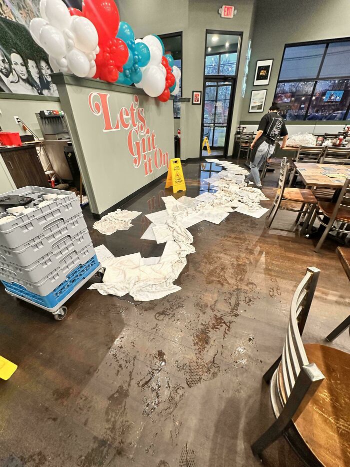 A restaurant employee cleans up a large water spill using towels and a wet floor sign, showing service industry workers challenges.