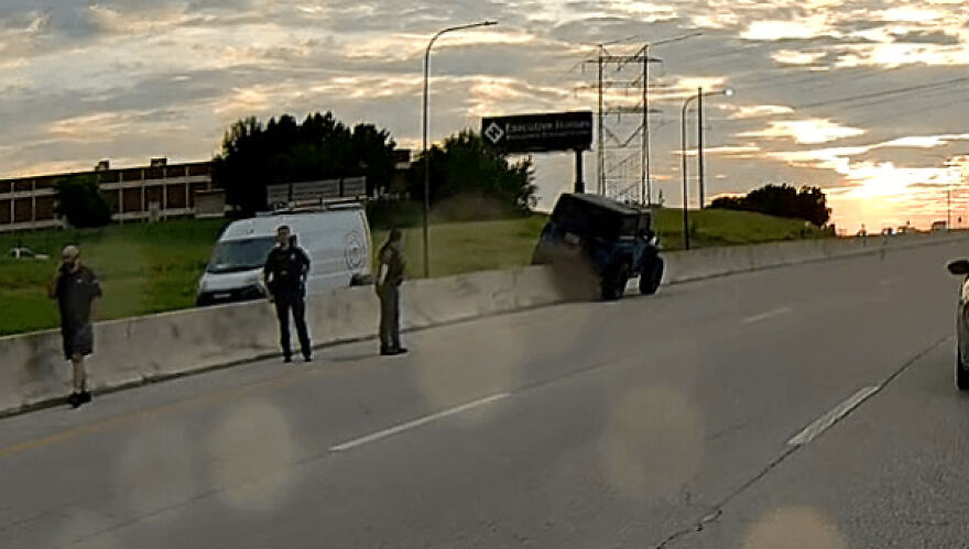 A dark blue Jeep crashed against a highway barrier with officials nearby, indicating a rough day behind the wheel.