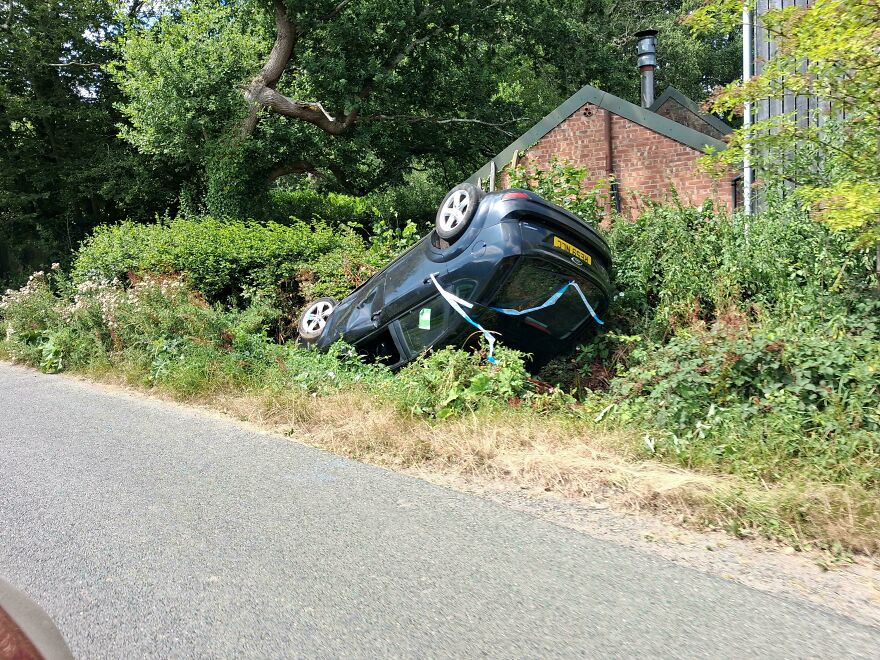A black car lies overturned in a ditch next to a road, showing a rough day behind the wheel.