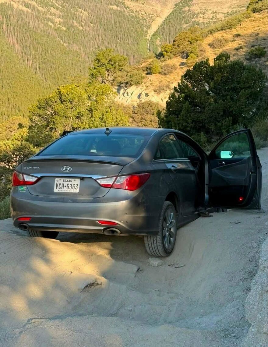 A dark gray sedan parked precariously on a dirt road on a steep incline, demonstrating a rough day behind the wheel.