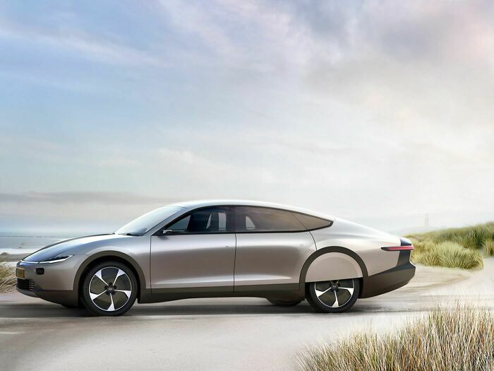 A futuristic silver car, representing ingenious technologies, parked on a sandy beach with tall grass and a lighthouse in the distance.
