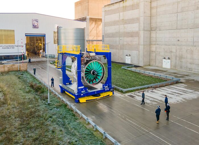 A massive Rolls-Royce aircraft engine, an example of genius technologies, on a transport rig outside a facility with workers nearby.
