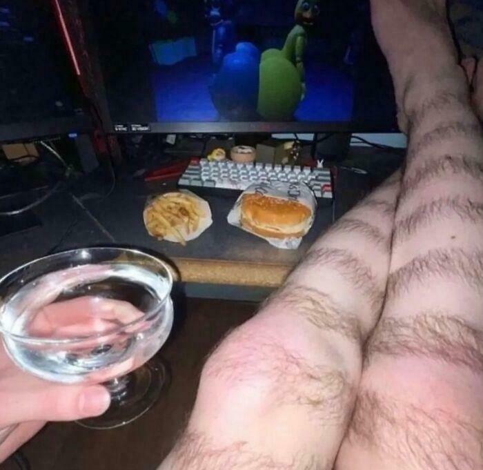 Hairy legs next to a computer screen showing a video game, with fast food and a drink nearby, a confusing image.