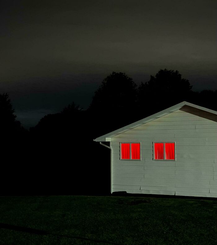 A confusing image of a small white house with two glowing red windows against a dark, treelined background, needing a priest.