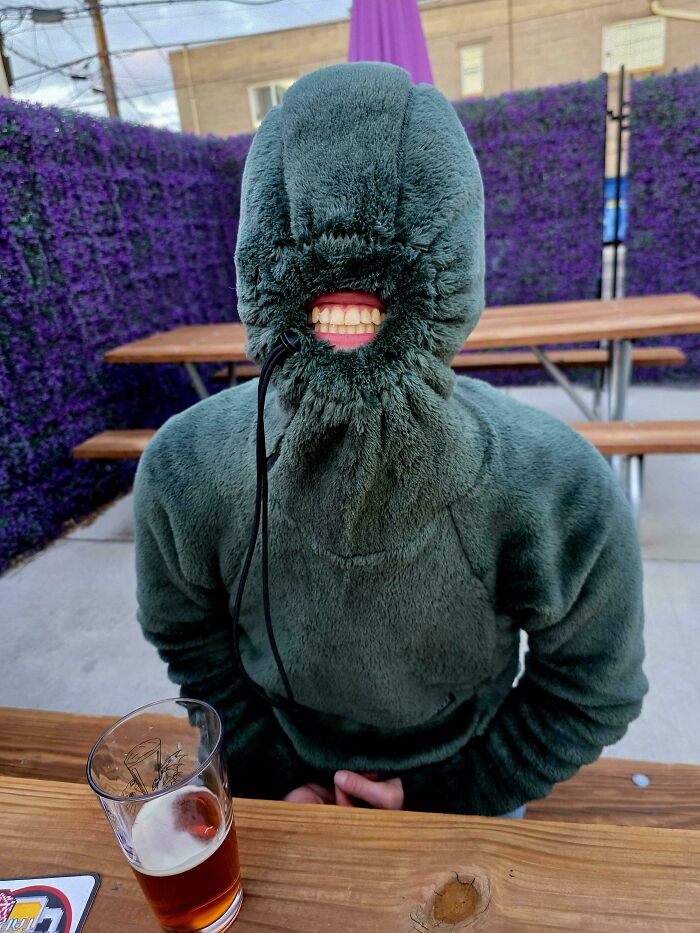 A creepy image of a person wearing a green hoodie with only fake teeth visible, sitting at a table with a beer, needing a priest.