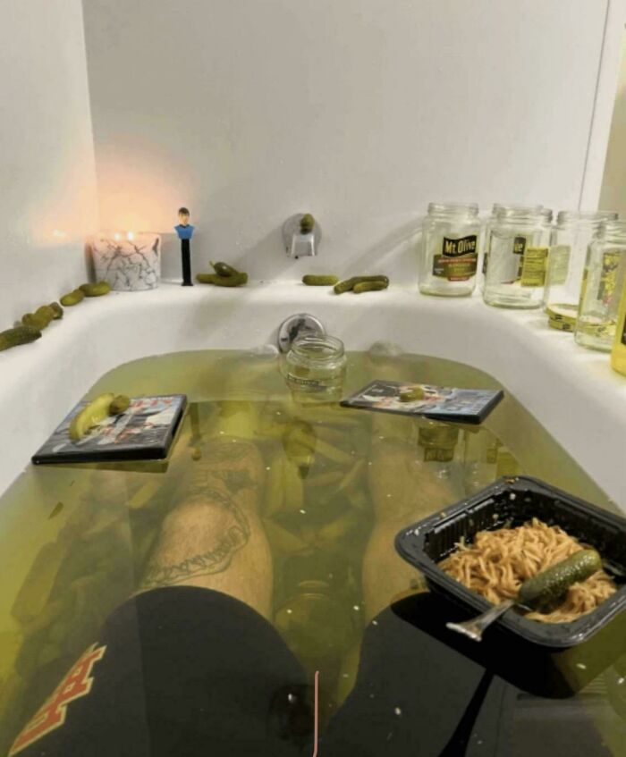 A person soaking in a bathtub filled with pickles and murky green water, with noodles and more pickles on the side, a truly creepy and confusing image.