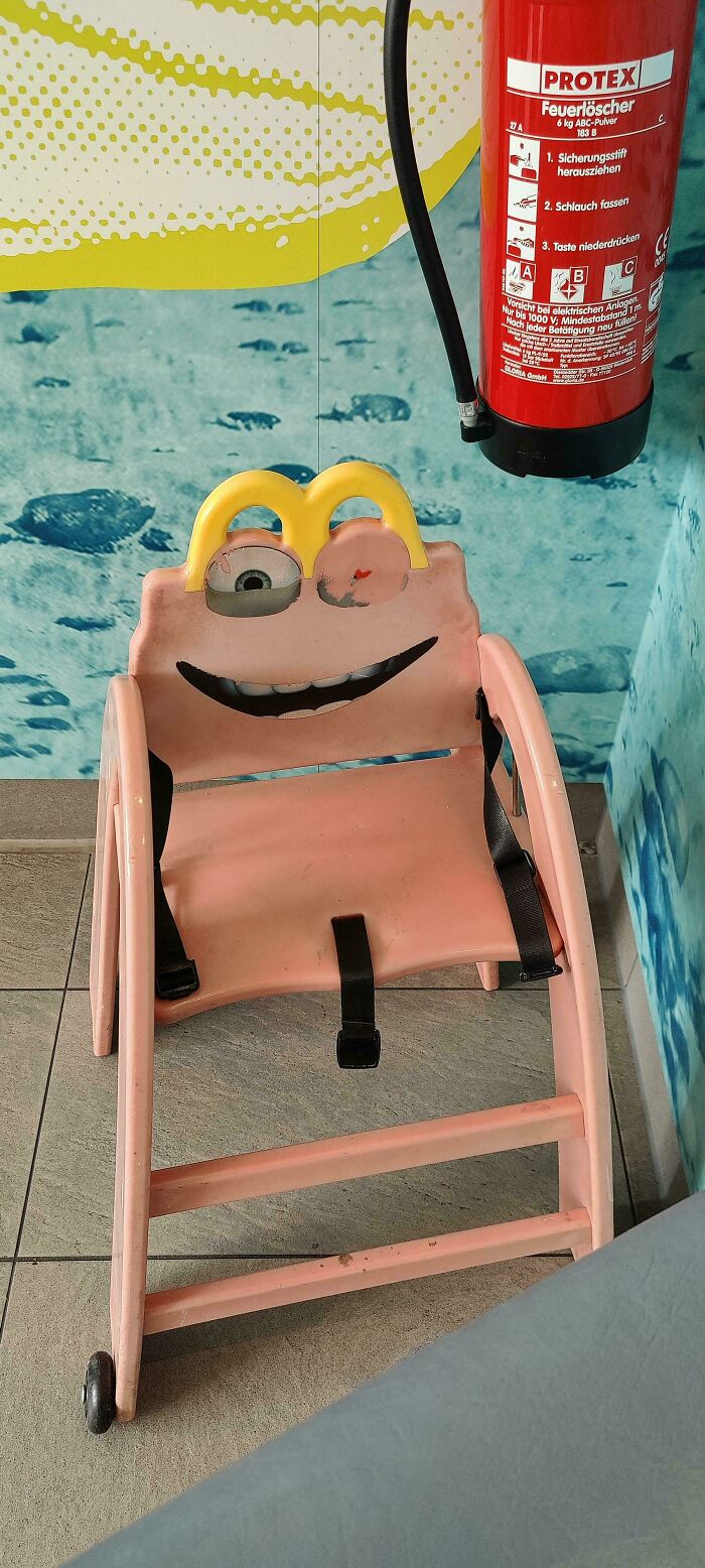 A creepy and confusing image of a pink child's chair with a frog-like face and black straps, needing a priest.