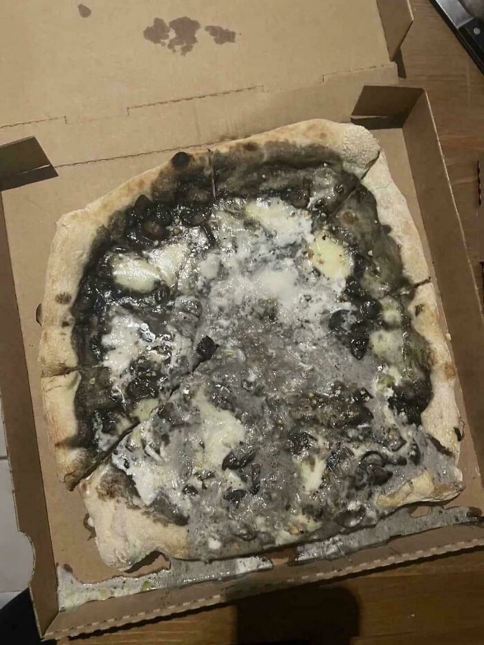 A creepy and confusing image of a black and white pizza with a strange, possibly moldy, topping in a box, needing a priest.