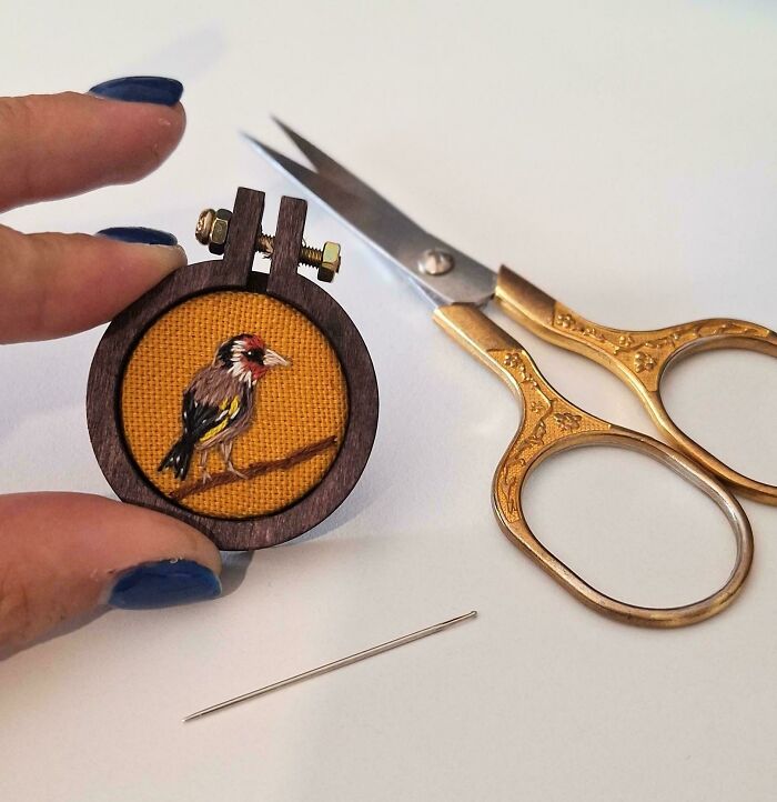A tiny hand holding a miniature bird embroidery project next to scissors