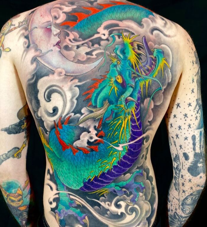 Full back tattoo art depicting a colorful dragon swirling around a crescent moon and clouds, an example of inspiring tattoo art.