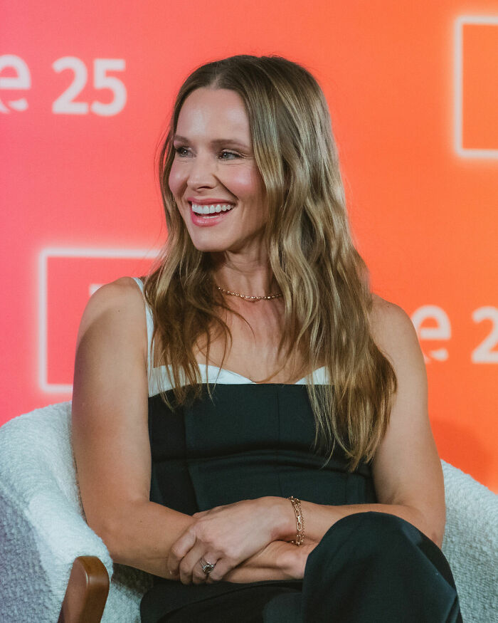 Kristen Bell career highlights, smiling and posing for a photo. She has long, wavy blonde hair and is wearing a black and white top.
