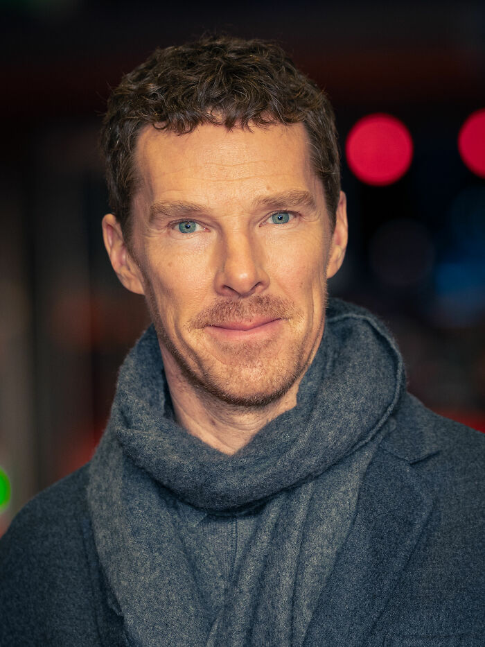 Benedict Cumberbatch close-up, showing his blue eyes and a subtle smile. He is wearing a dark grey coat and a matching scarf.
