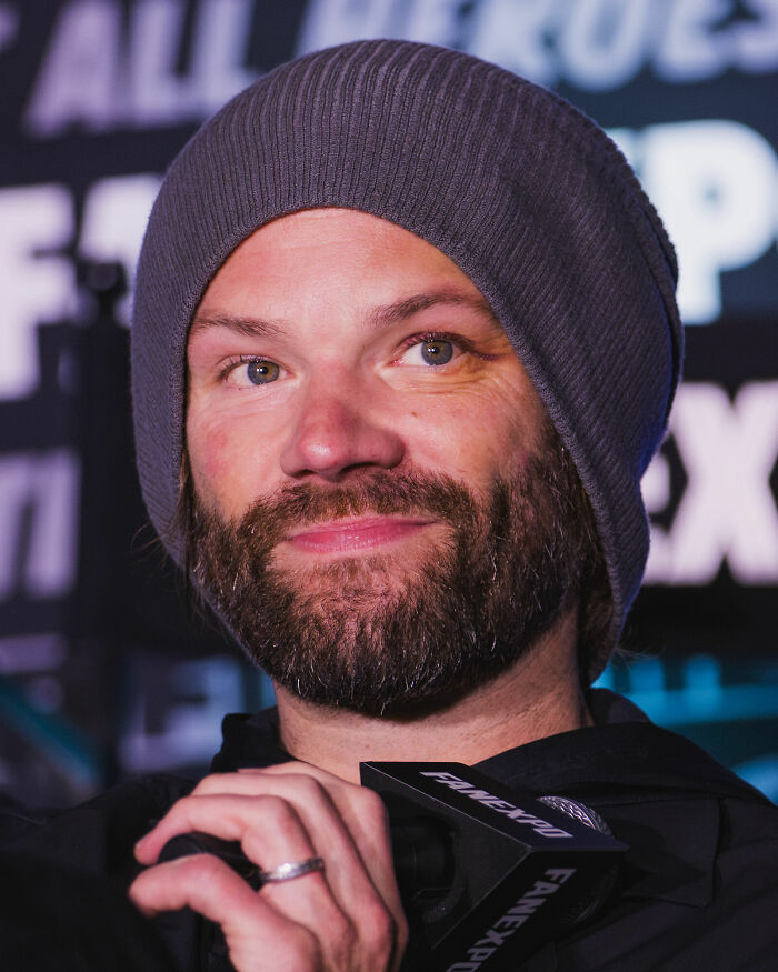 Jared Padalecki wearing a gray beanie, with a beard, holding a microphone, smiling faintly with bright eyes.